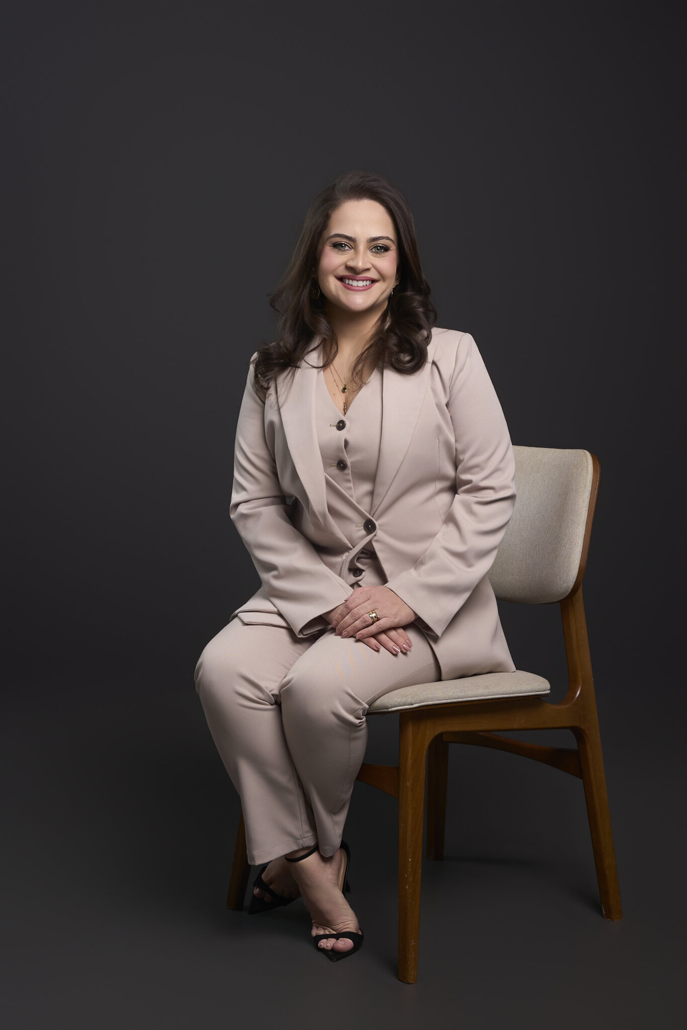 Dra. Claryssa Buriti sorrindo, sentada em cadeira de madeira clara, com terno bege claro